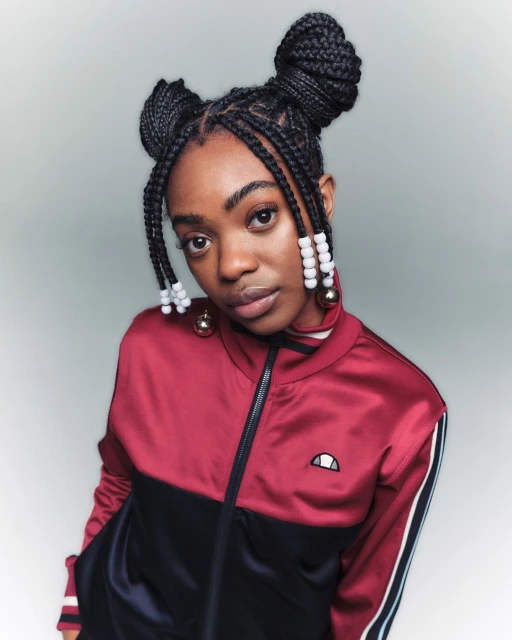Portrait of a young woman with braided hair styled in space buns, wearing a burgundy and black track jacket in a studio setting.