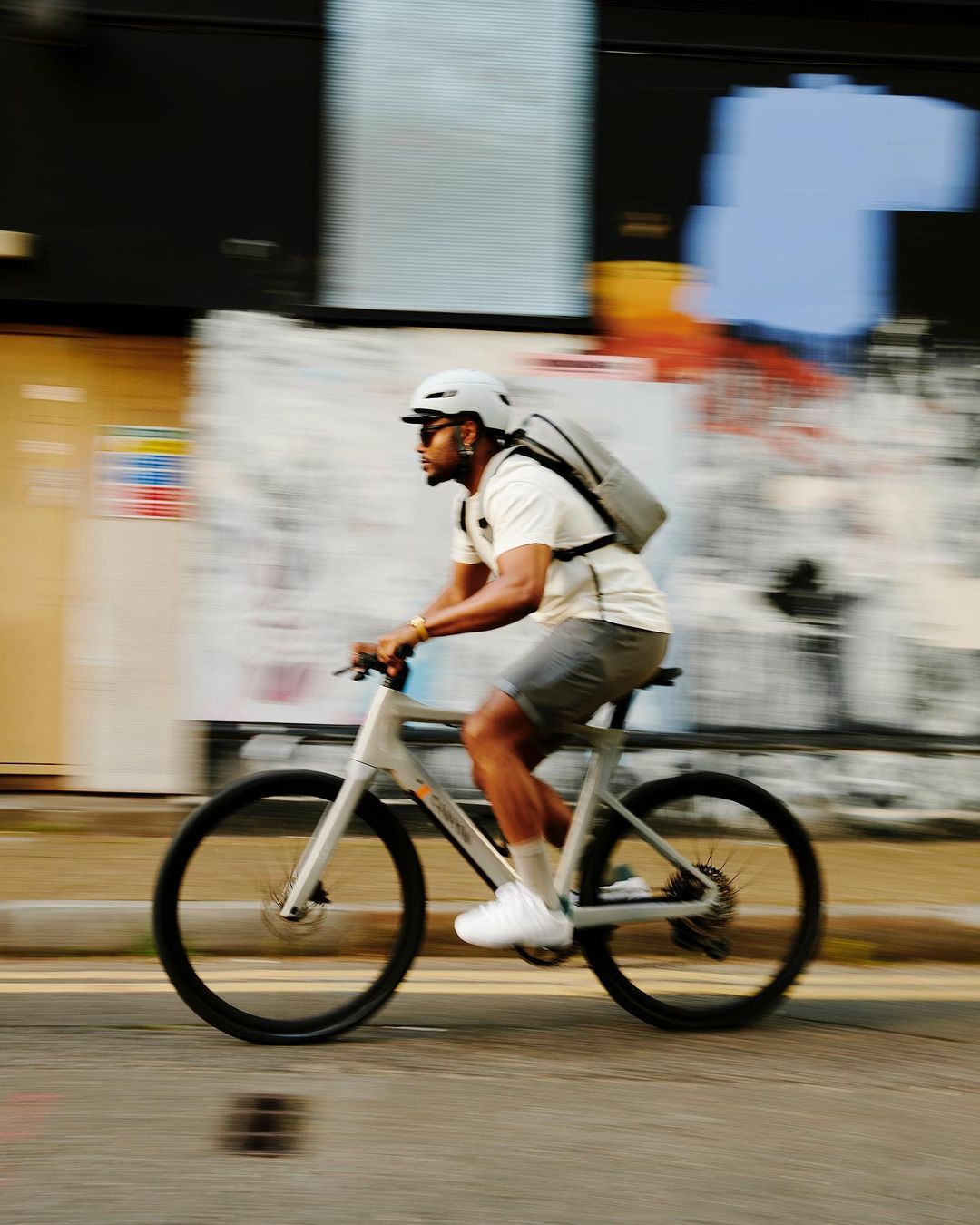 An adult male rides an electric bicycle on a city street with a motion blur effect, wearing a helmet, backpack, and casual clothing.