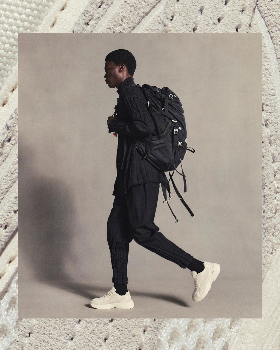 Side profile of a black male model wearing a black outfit and a black backpack while walking in a photo studio with white sneakers.