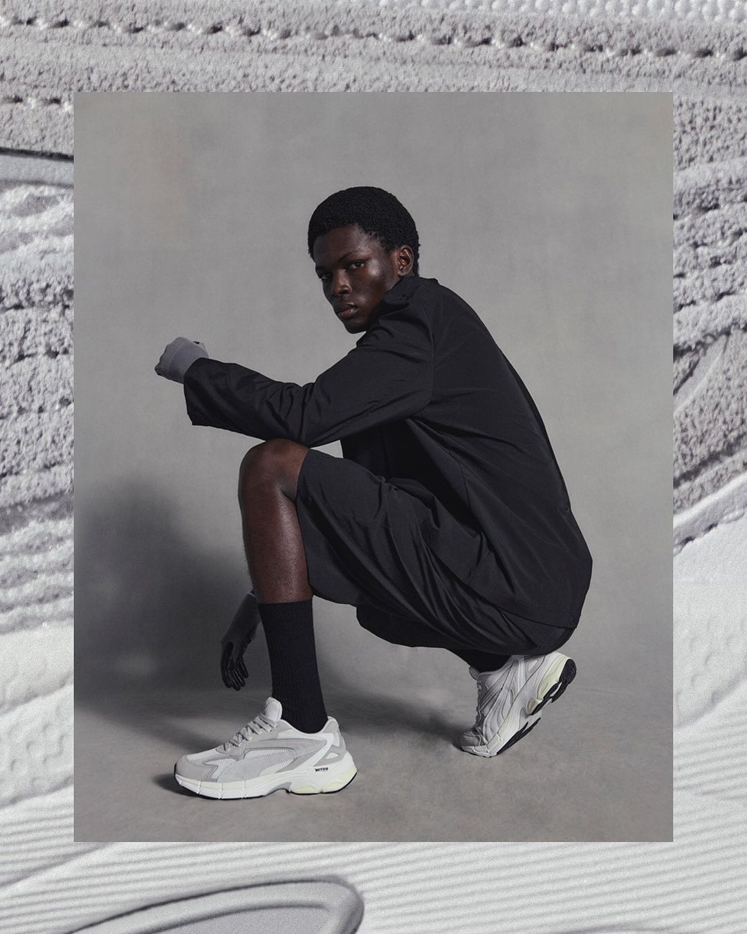 A Black male model poses in a crouch wearing black clothing and off-white sneakers, set against a grey backdrop with textural foreground.