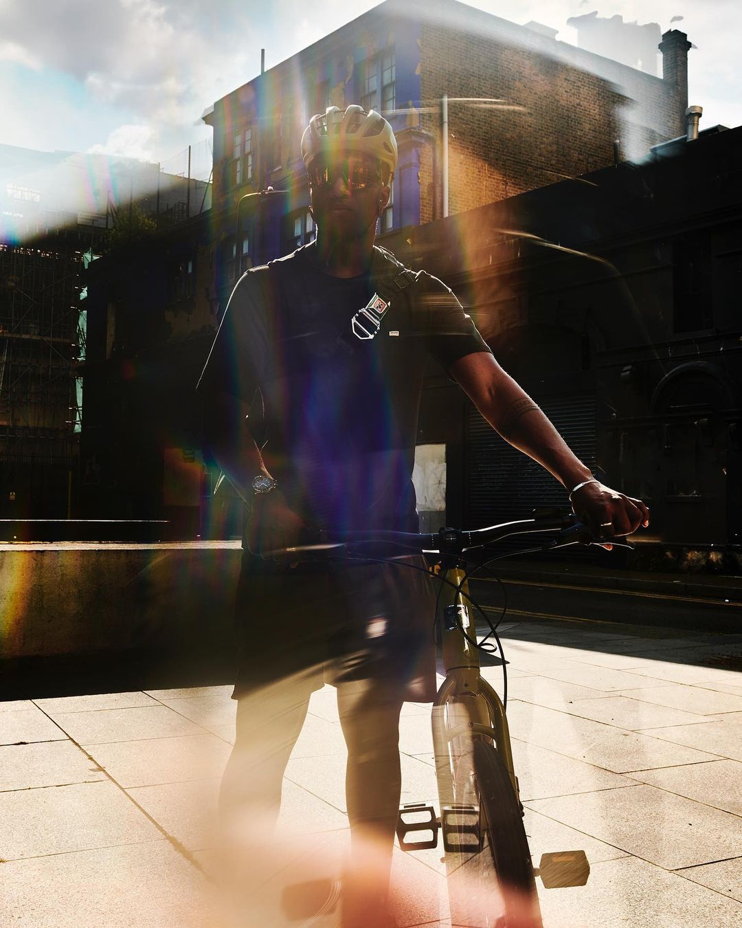 Cyclist pauses on a bike in sunlight, wearing helmet and dark athletic wear in an urban area, buildings faintly in the background.