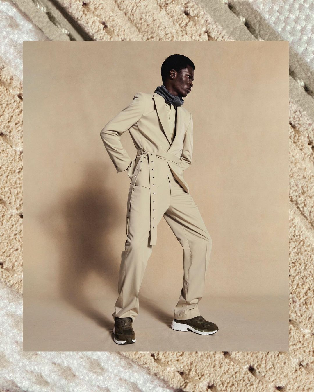An adult Black male poses in a studio wearing a light tan suit, dark shoes, and a dark gray neck covering, with a soft tan background.