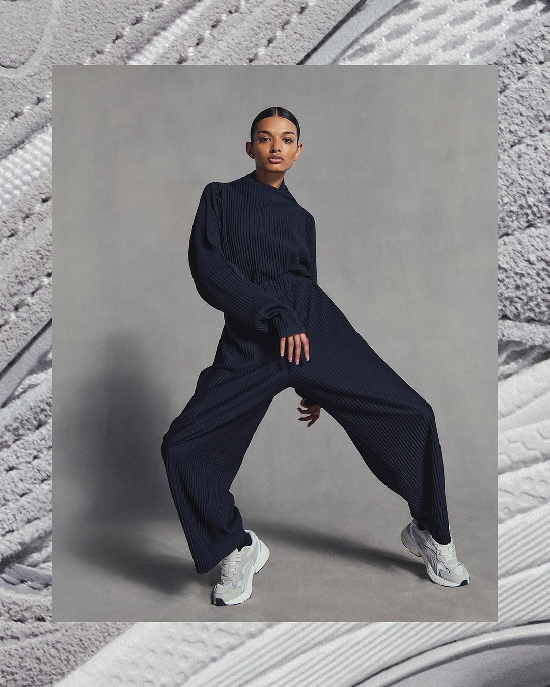 An adult female model poses in a blue pleated jumpsuit and white running shoes against a grey background, framed by a textured border.
