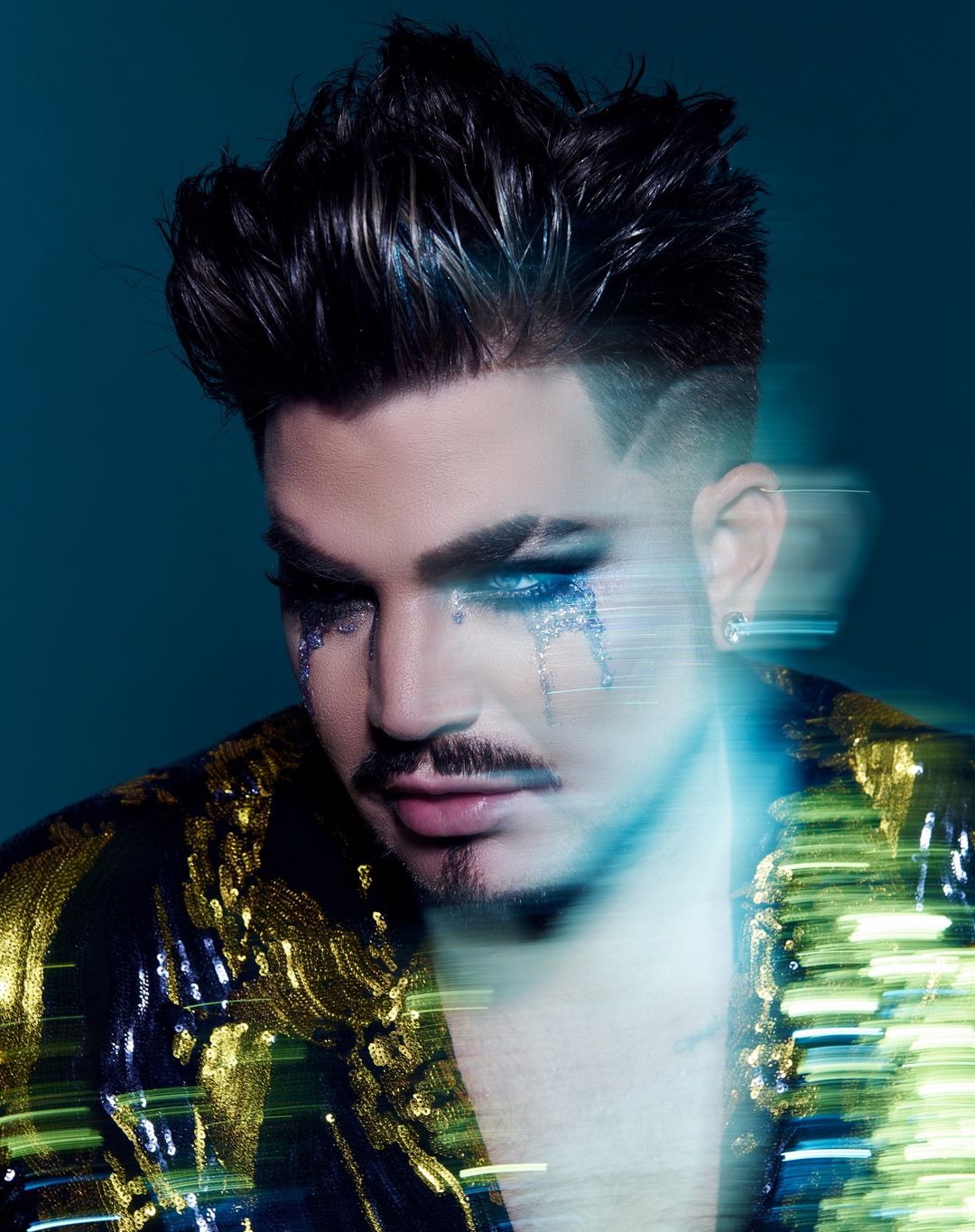 Close-up portrait of a man with styled dark hair, wearing a gold and black jacket with glitter makeup under his eyes.