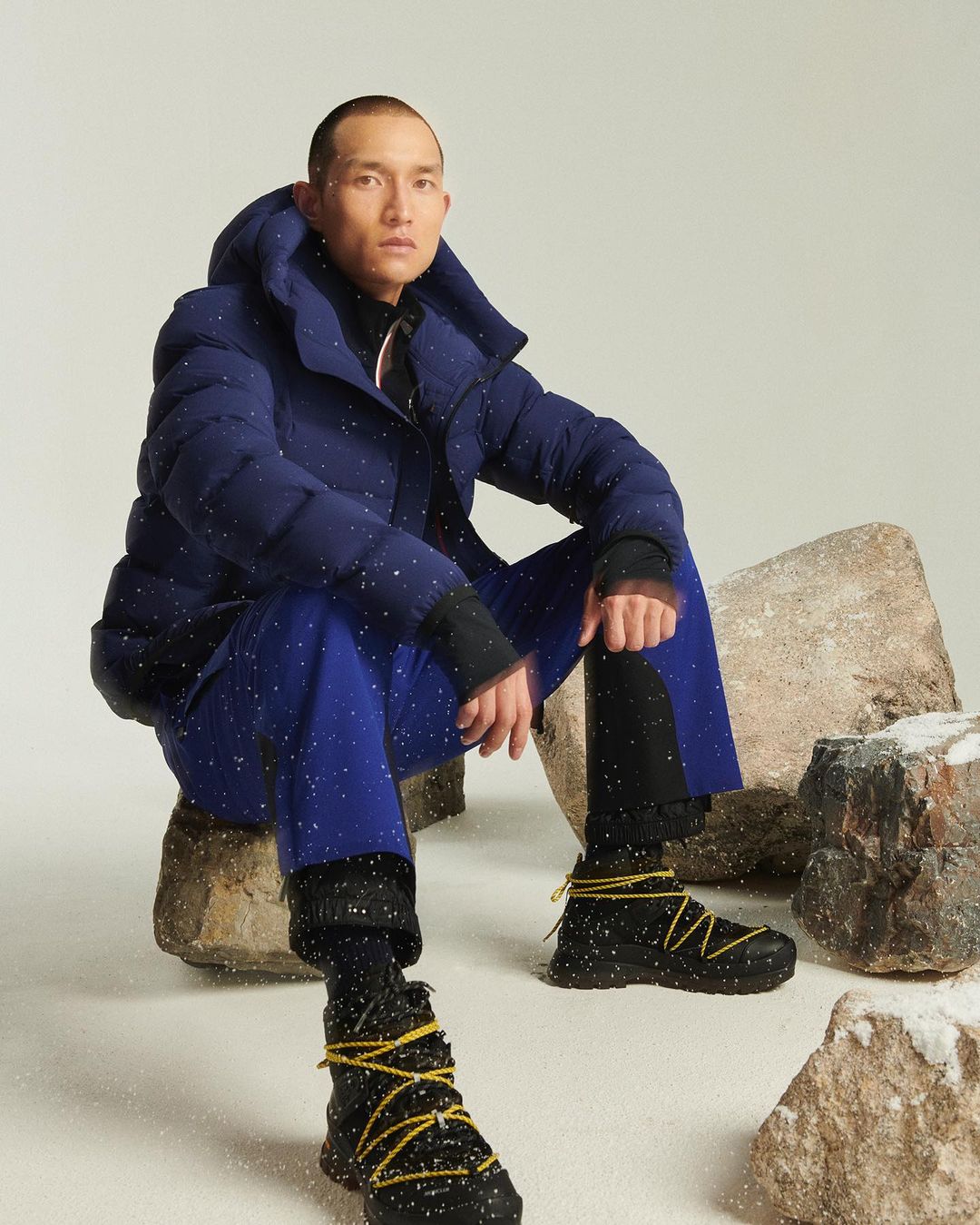 A man in a blue puffer jacket and blue pants sits on rocks with snowy footwear in a staged photo studio setting with artificial snow.