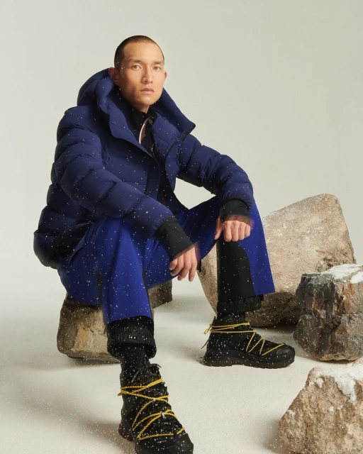 A man in a blue puffer jacket and blue pants sits on rocks with snowy footwear in a staged photo studio setting with artificial snow.