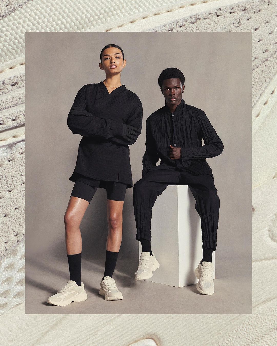 Two fashion models, an adult woman and man, pose indoors wearing black clothing and off-white sneakers. Neutral studio background.