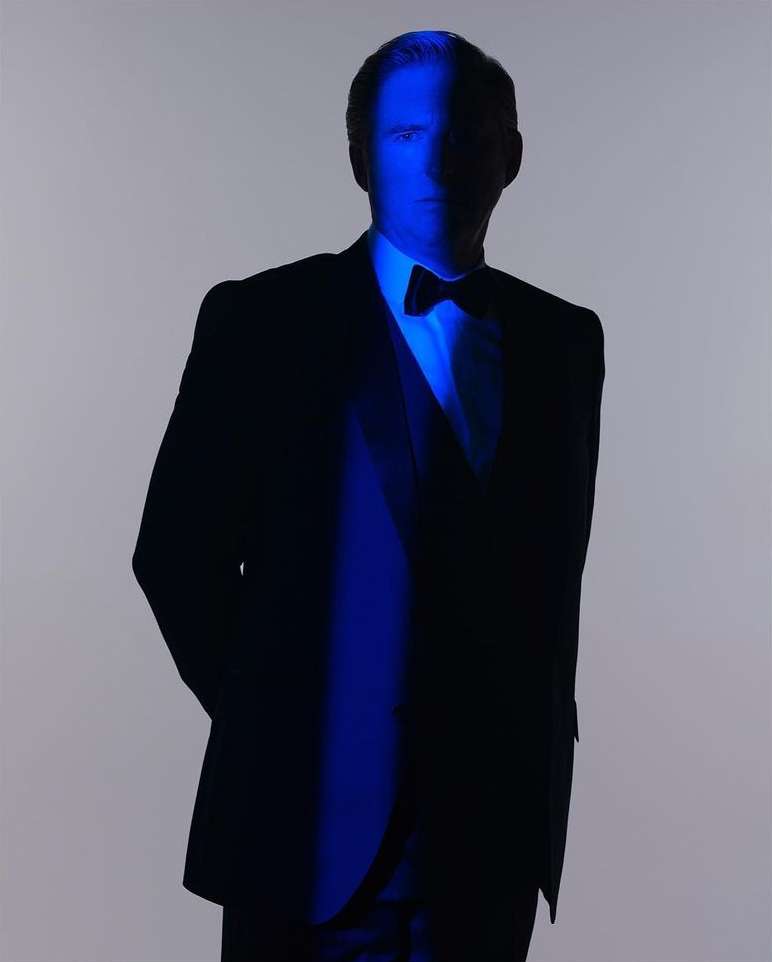 A man in a black suit and bow tie stands against a light background, highlighted by a vertical beam of blue light on his face.