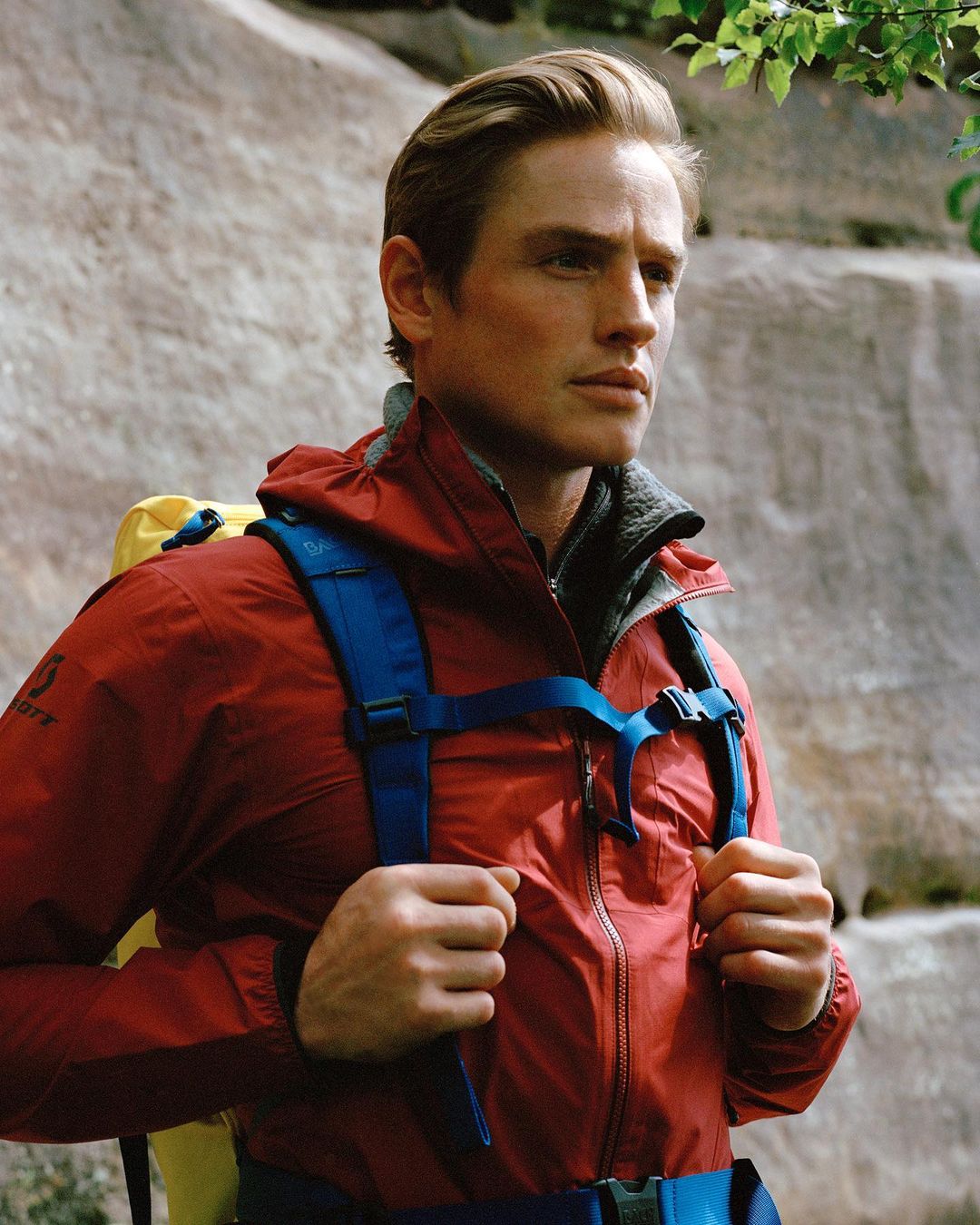 Man in a red jacket with a backpack hiking in a natural environment with rock formations in the background and green leaves.