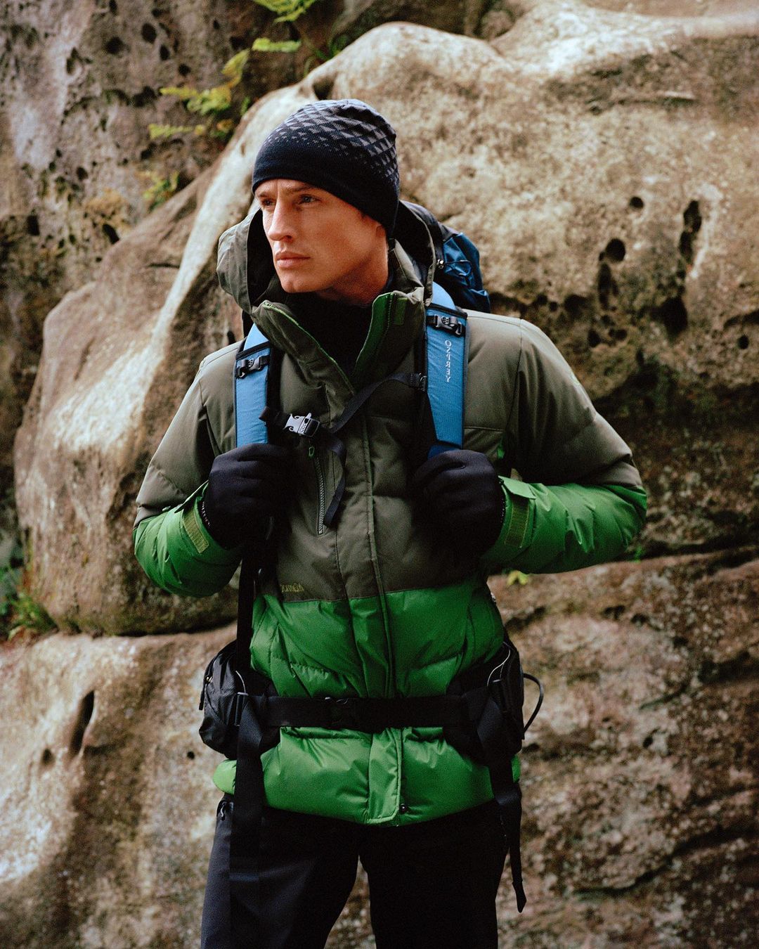 Man wearing a green and grey jacket, backpack, and knit hat stands in front of large rock formations in an outdoor setting.