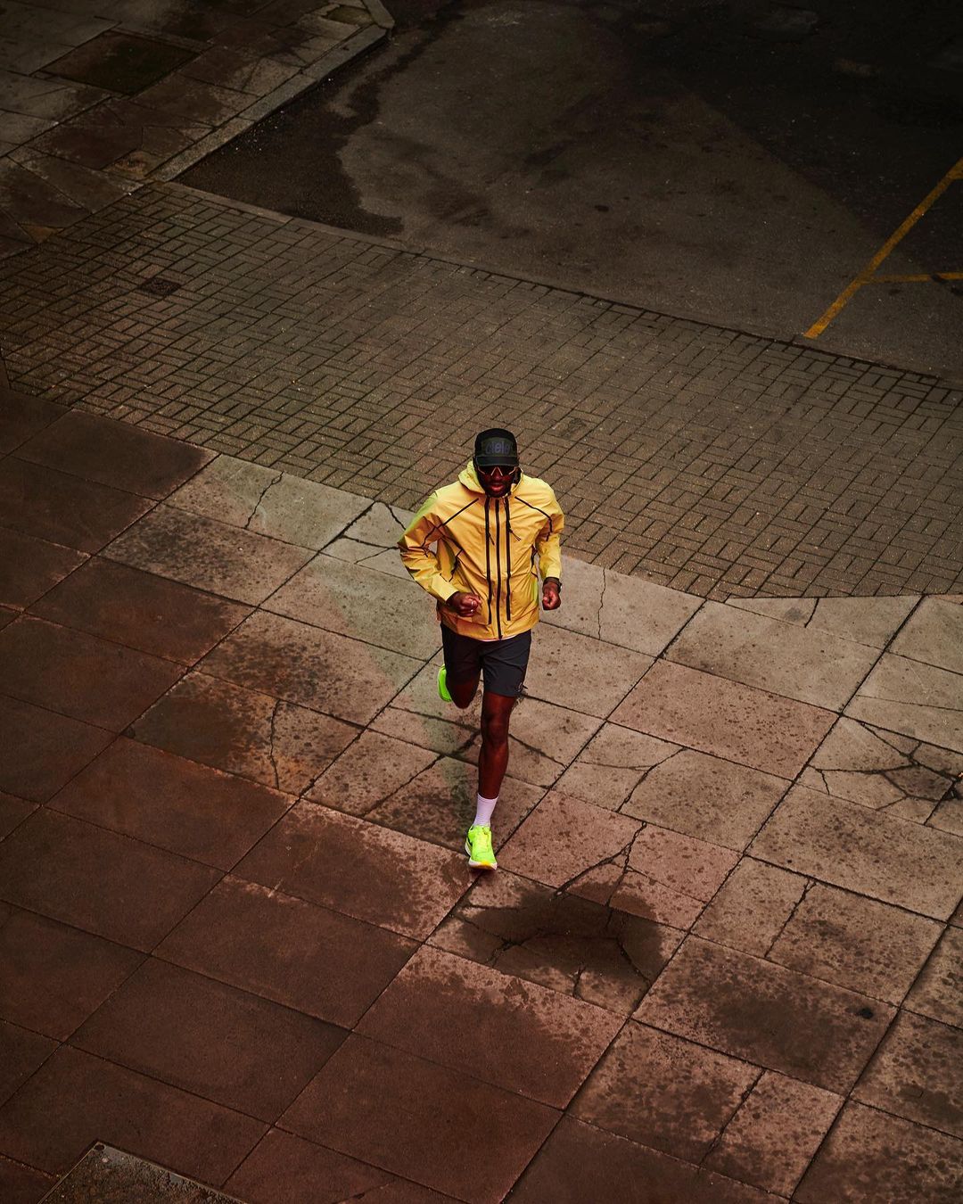 Overhead shot of a man running on a brick sidewalk in yellow windbreaker, shorts, and neon running shoes, creating a sense of motion and urban fitness.