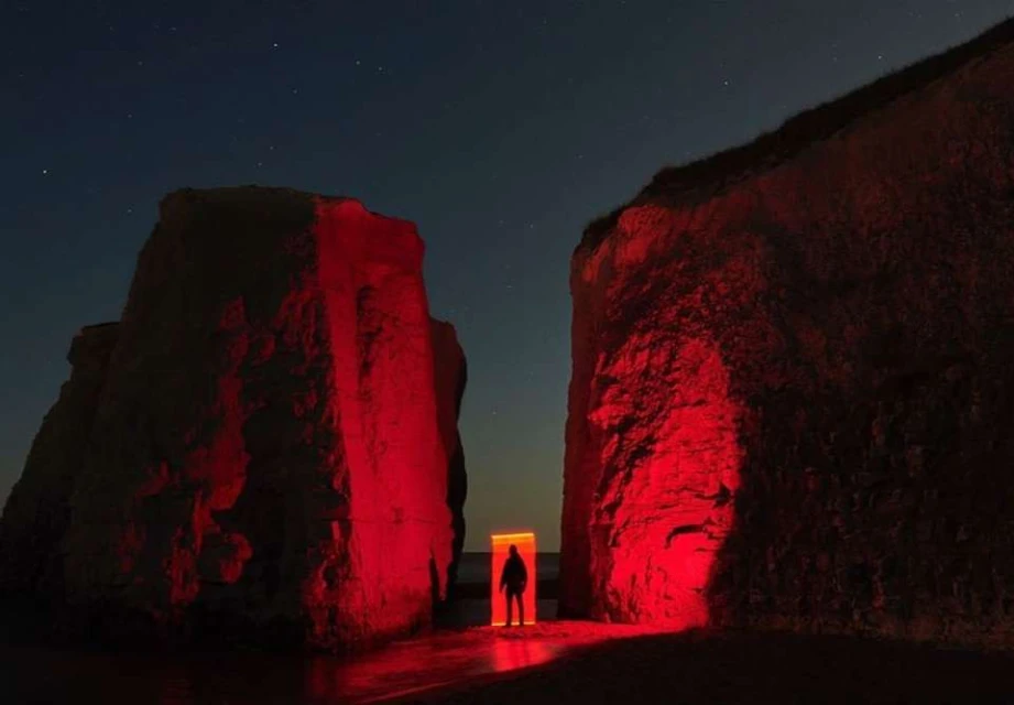 Silhouette of a person standing in a red-lit doorway between two large rock formations on a beach under a night sky with stars.
