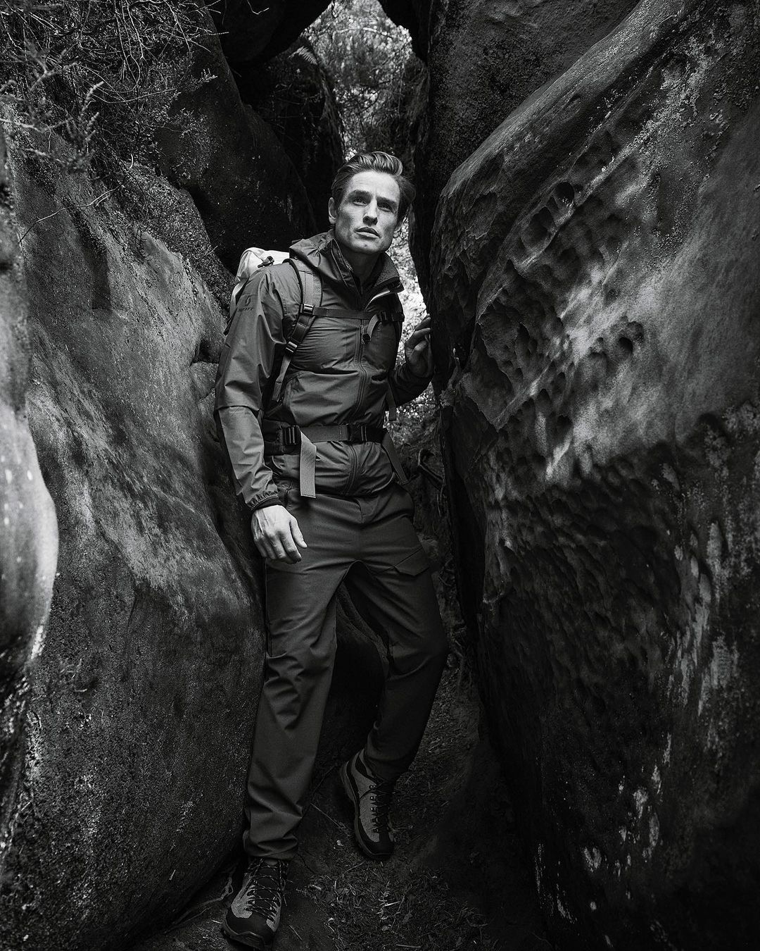 Monochrome portrait of a man in a hiking jacket and backpack standing between rock formations in a wilderness area.
