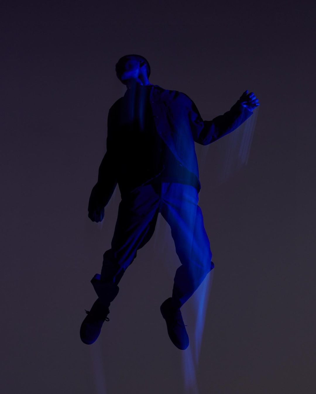 An adult poses mid-air, bathed in deep blue lighting. The individual wears dark clothing against a muted gray background, creating a surreal effect.