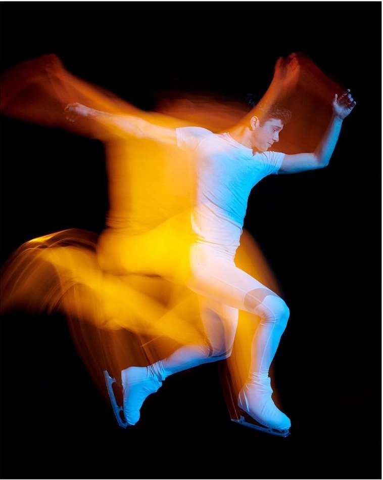 A male figure skater in motion against a black background, captured with intentional blur to emphasize the speed and fluidity of the performance.