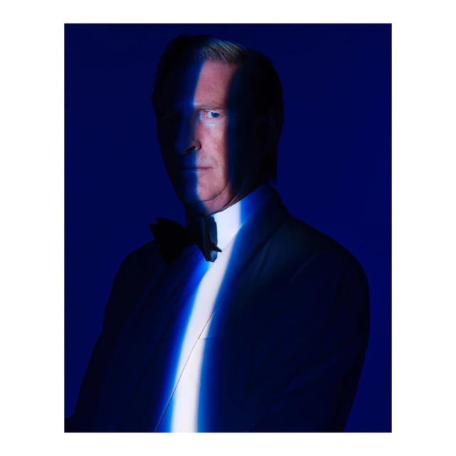 Portrait of an adult male with auburn hair wearing a suit with a bow tie against a dark blue background with a beam of light on face.