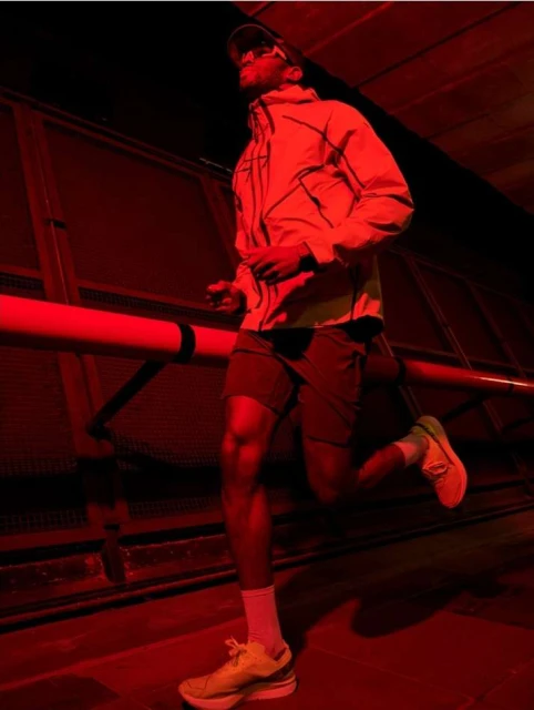 Man in windbreaker and running shoes running on elevated walkway under red lighting at night.