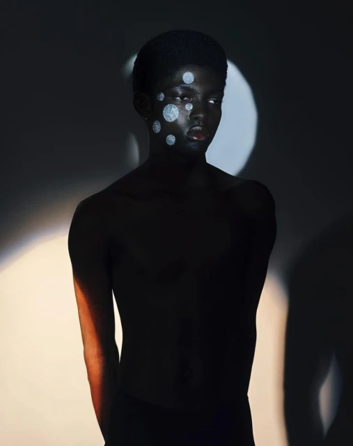 Portrait of a young black man with silver circles painted on his face, in front of a grey background with soft, diffused lighting.