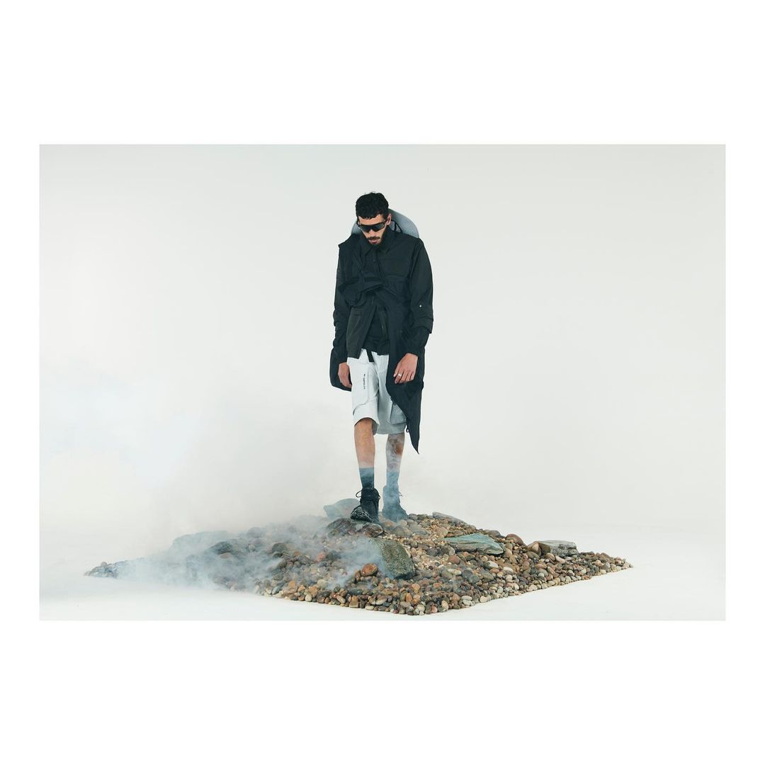 A model in sunglasses, a black jacket, and white shorts stands on rocks surrounded by smoke in a studio fashion shot.