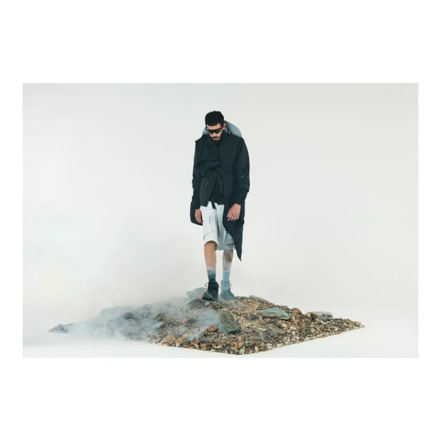 A model in sunglasses, a black jacket, and white shorts stands on rocks surrounded by smoke in a studio fashion shot.
