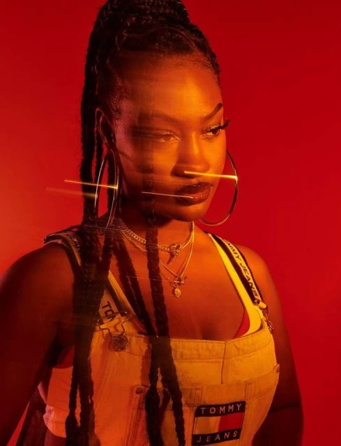 Eye-level portrait of a young Black woman with braided hair wearing denim overalls against a red background, illuminated by soft lighting.