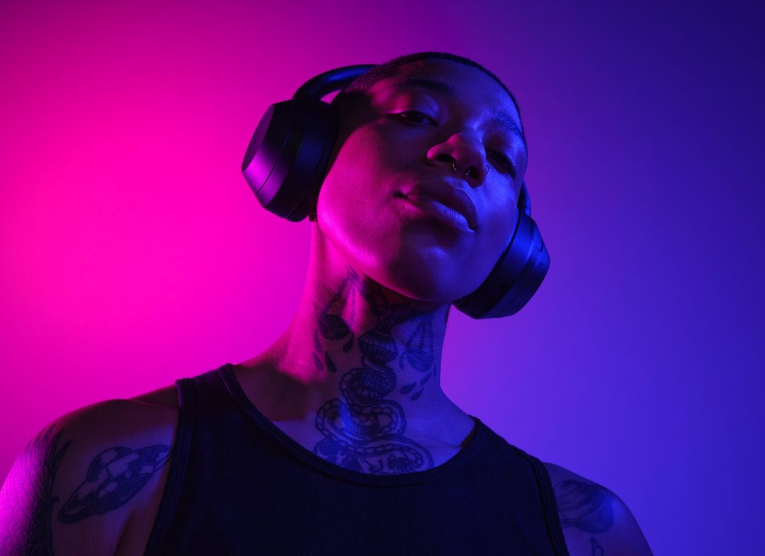 Adult individual with wireless headphones. Tattoos visible on the neck and shoulders. Pink and blue gradient background lighting.