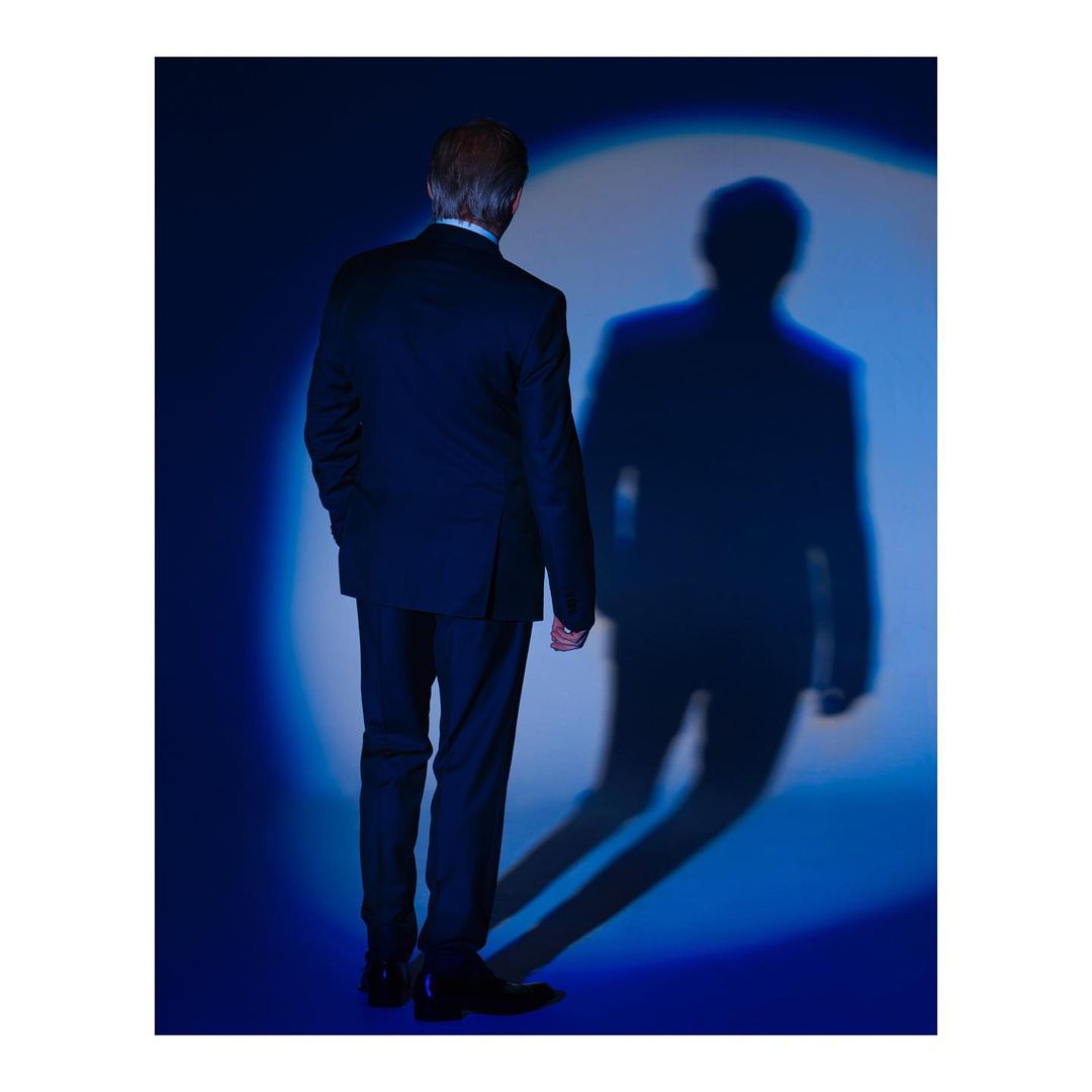 Man in dark suit viewed from behind standing against a blue wall with a circular spotlight projecting a sharp shadow.