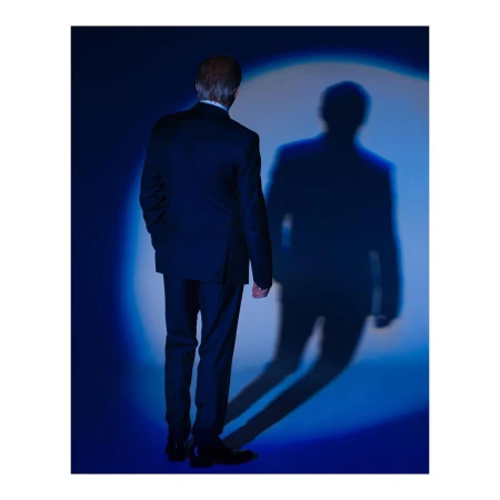 Man in dark suit viewed from behind standing against a blue wall with a circular spotlight projecting a sharp shadow.