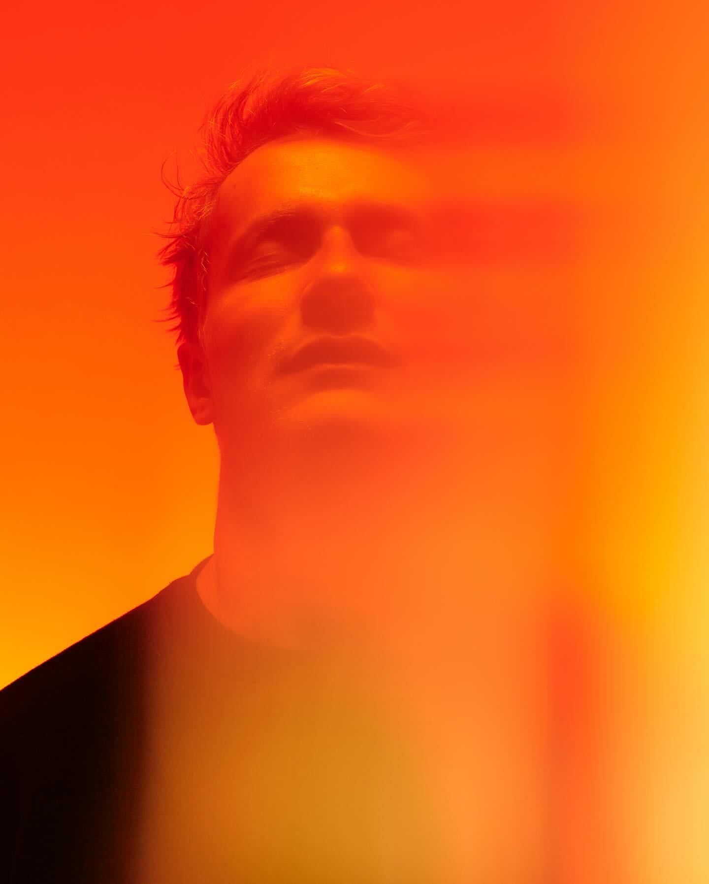 Close-up studio portrait of an adult male looking up with eyes closed, lit with vibrant red and orange light in a warm, diffused glow.