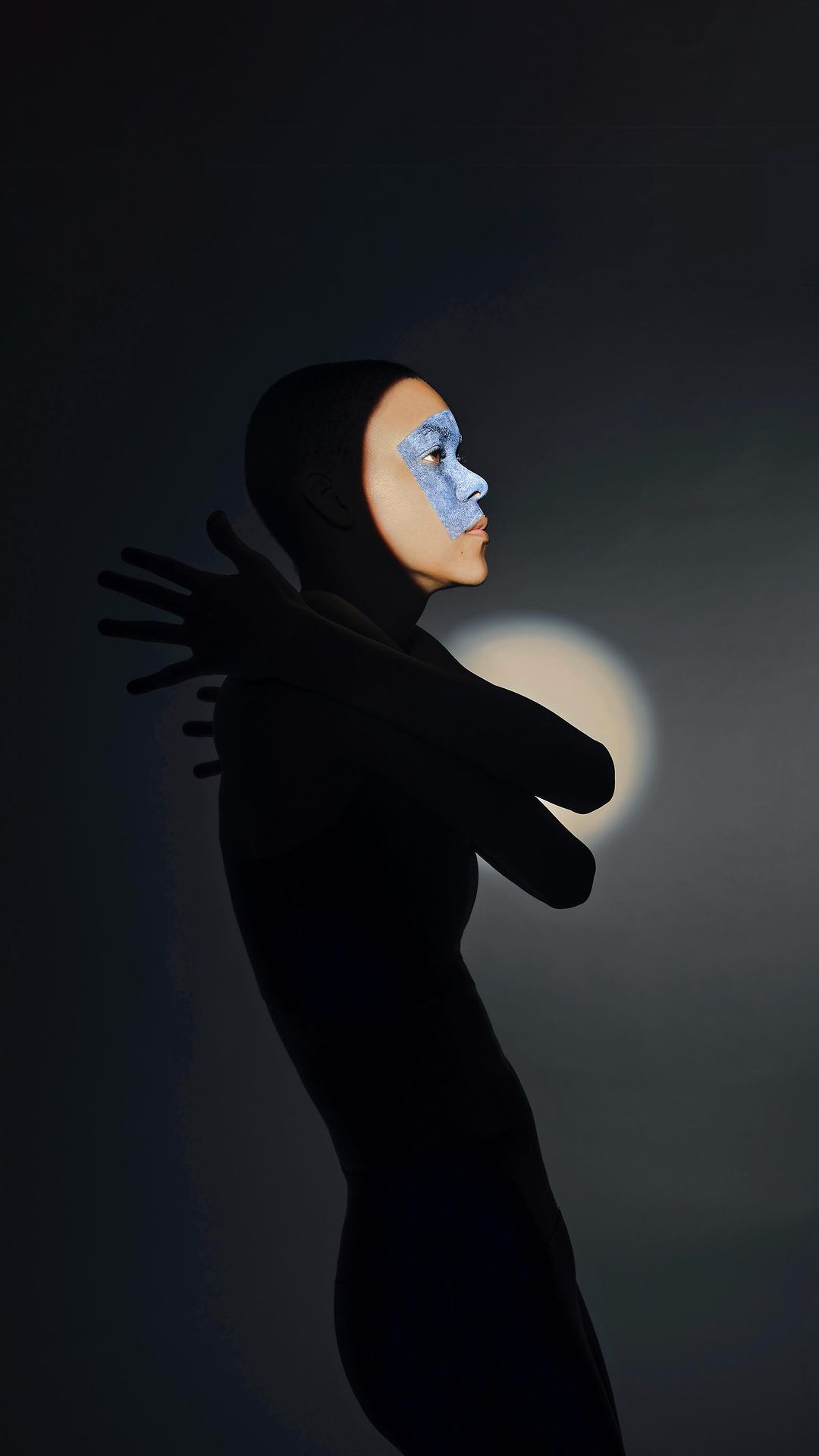Portrait of a person in a hooded garment, face painted with blue makeup, side lit against a dark grey background with hands gently folded.
