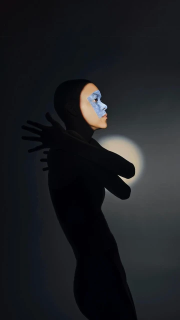 Portrait of a person in a hooded garment, face painted with blue makeup, side lit against a dark grey background with hands gently folded.