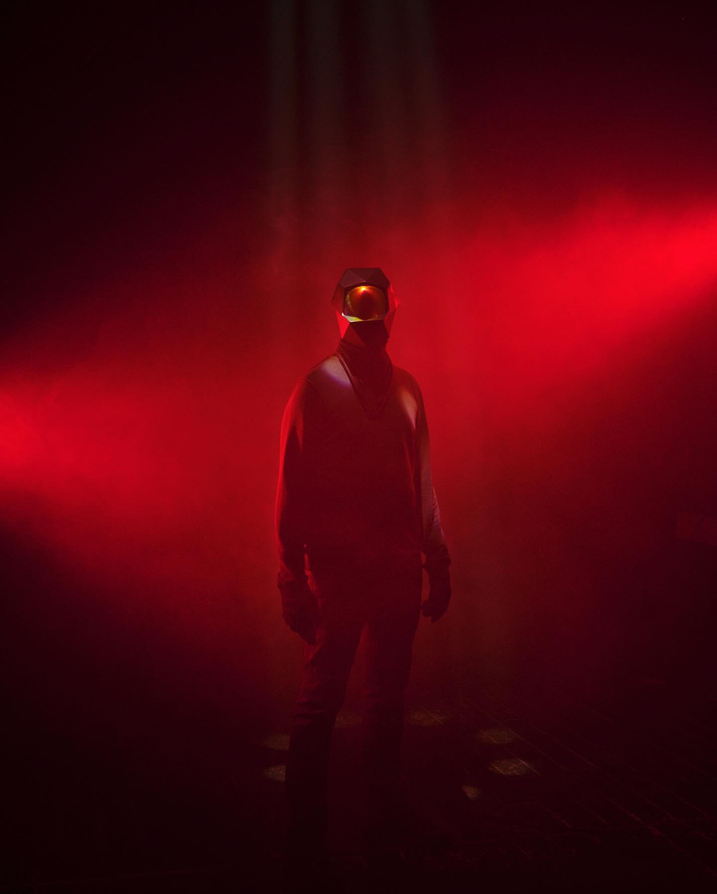 Figure cloaked in dark clothing and goggles stands in a foggy, reddish-lit atmosphere, with the light creating an eerie effect.