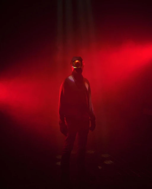 Figure cloaked in dark clothing and goggles stands in a foggy, reddish-lit atmosphere, with the light creating an eerie effect.