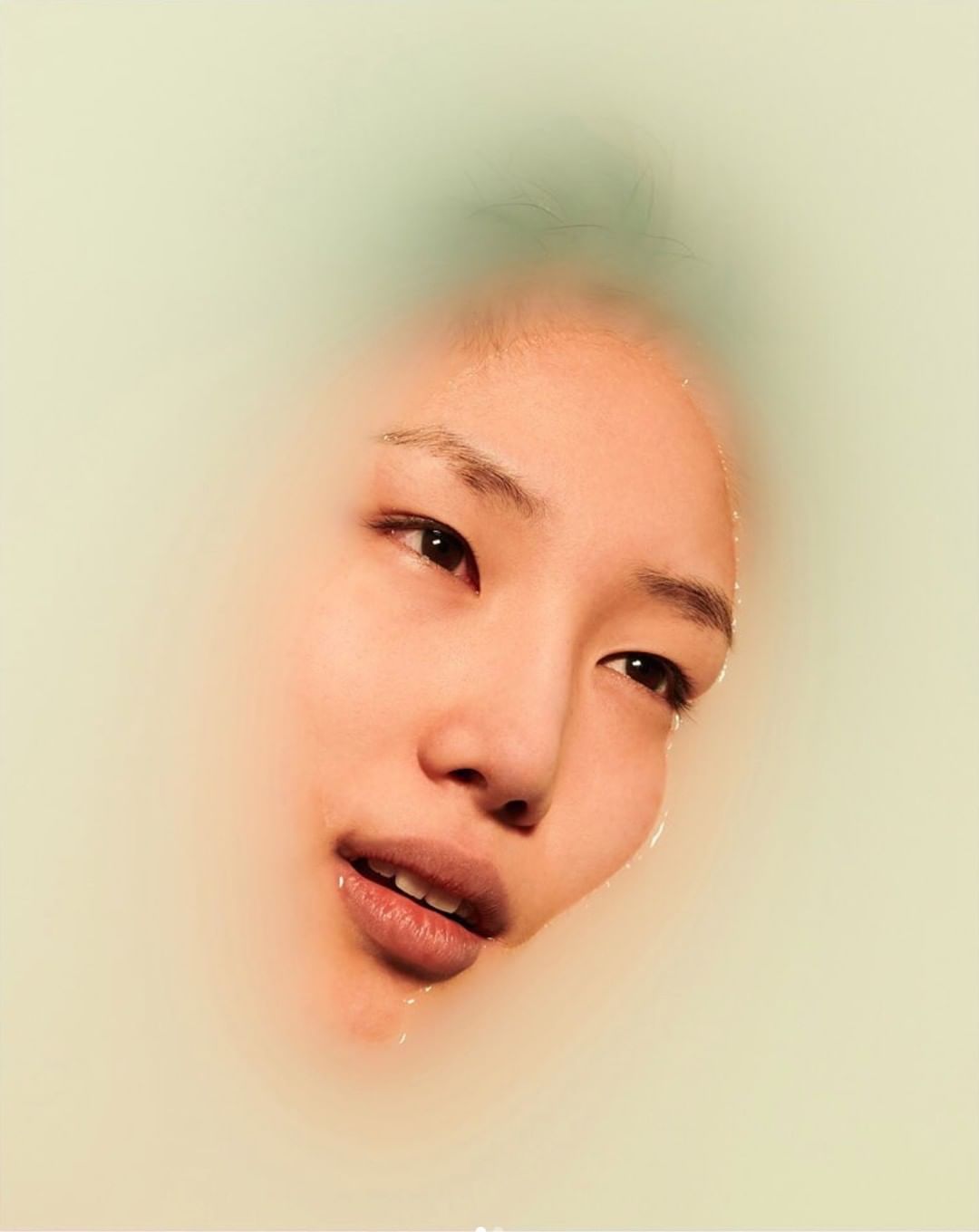 Asian woman's face emerging from milky water, eyes open, lips slightly parted, soft diffused light, beauty and skincare visual.