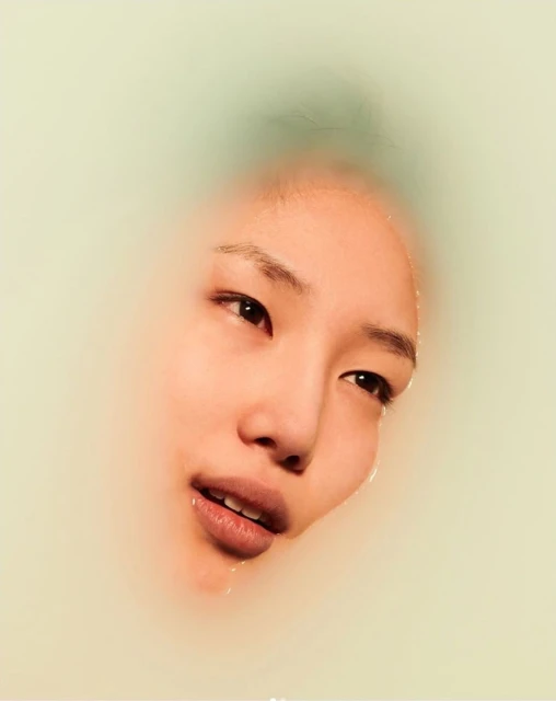 Asian woman's face emerging from milky water, eyes open, lips slightly parted, soft diffused light, beauty and skincare visual.