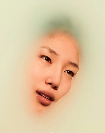 Asian woman's face emerging from milky water, eyes open, lips slightly parted, soft diffused light, beauty and skincare visual.