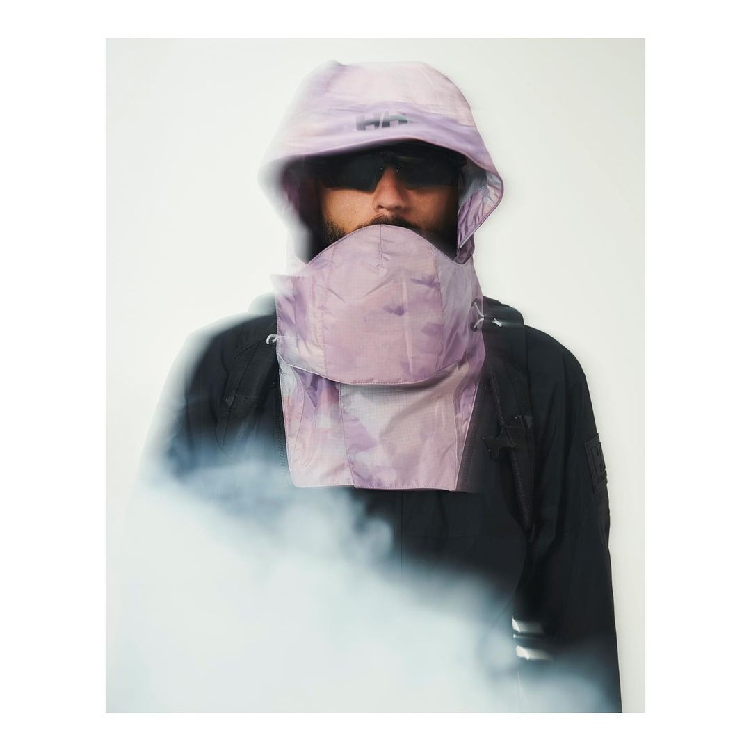 Portrait of adult male wearing black jacket, lavender hood, and sunglasses in a brightly lit photo studio with some artificial smoke.