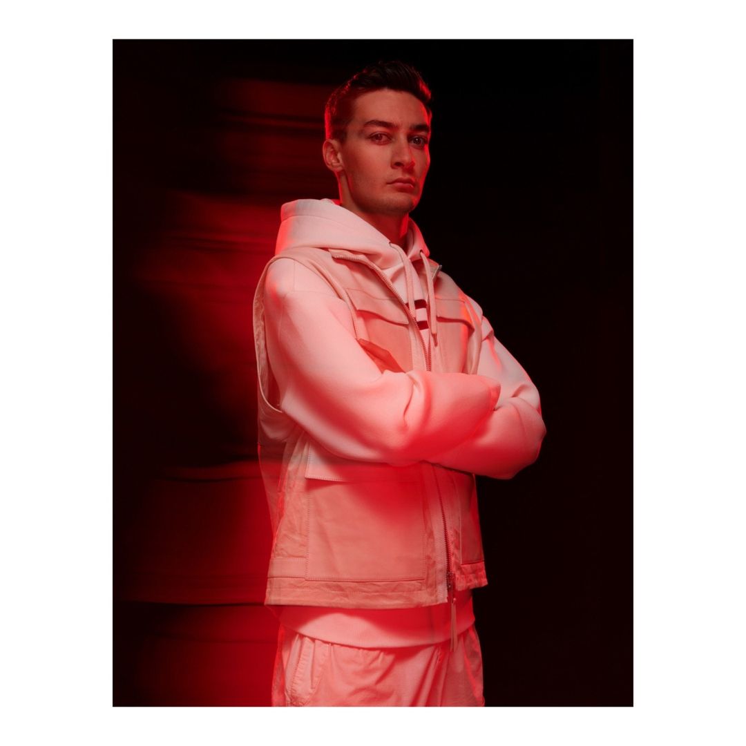 Portrait of a man wearing a white hoodie and vest with arms crossed under red lighting.
