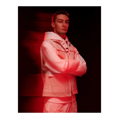 Portrait of a man wearing a white hoodie and vest with arms crossed under red lighting.
