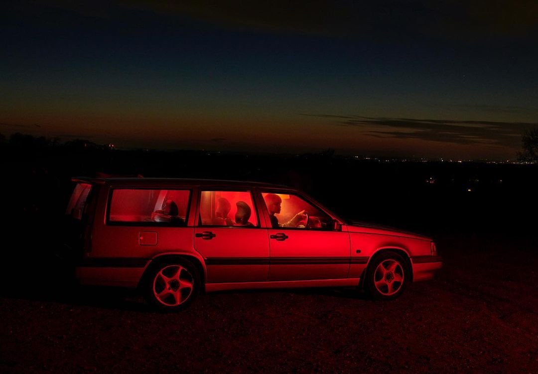 Car interior with three people illuminated in red light at night, outdoor dark landscape, atmospheric mood, roadside vehicle shot.