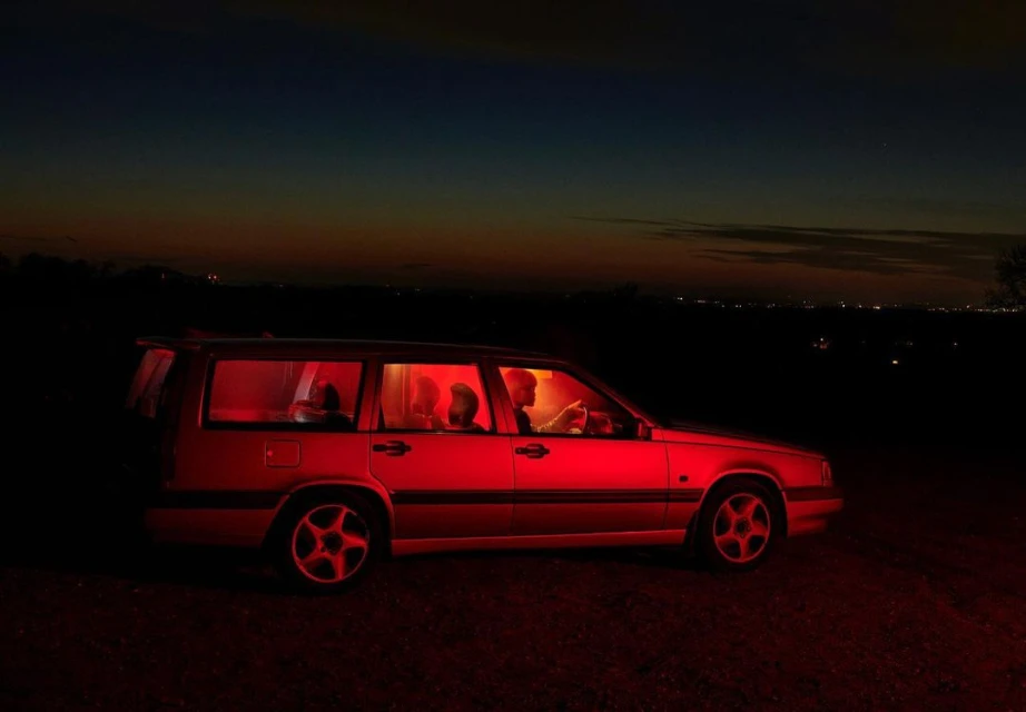 Car interior with three people illuminated in red light at night, outdoor dark landscape, atmospheric mood, roadside vehicle shot.