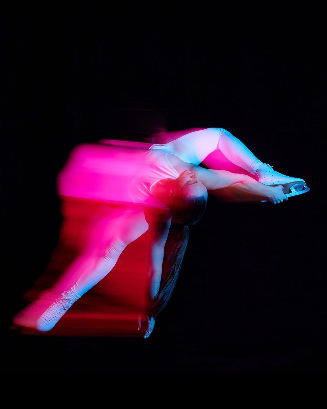 A person in white clothing and ice skates performing a skating move in a studio with pink and blue neon lighting.