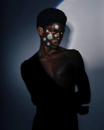 Portrait of a black man with silver glitter spots on his face and shoulder against a moody dark background.
