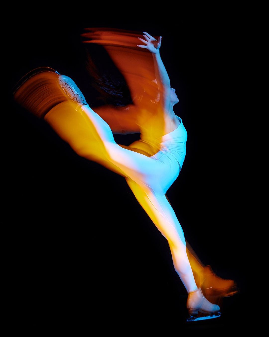 Dynamic long exposure shot of a female figure skater performing a Biellmann spin in a white bodysuit and skates against a black backdrop.