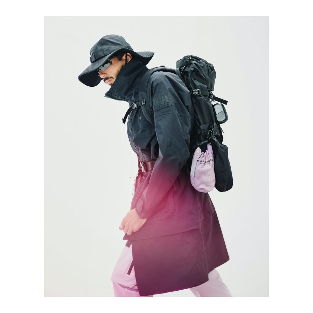 Man in dark coat and hat wearing backpack, walking in front of bright background. Fashion shot, studio lighting, outdoor-style clothes.