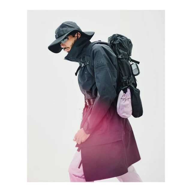 Man in dark coat and hat wearing backpack, walking in front of bright background. Fashion shot, studio lighting, outdoor-style clothes.