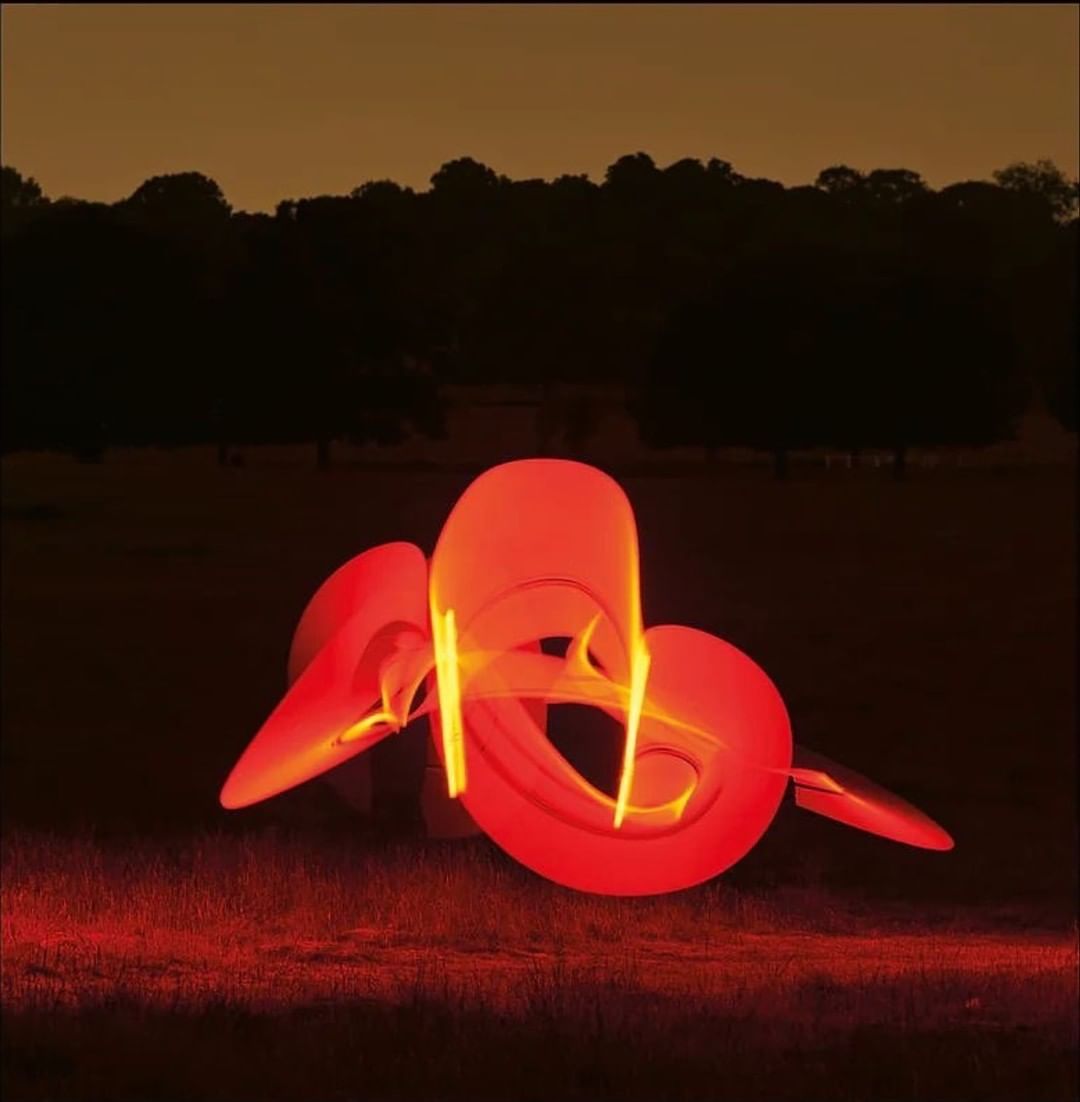 Abstract light painting in red and yellow glows over a grassy field with a dark line of trees under an orange sky at dusk.