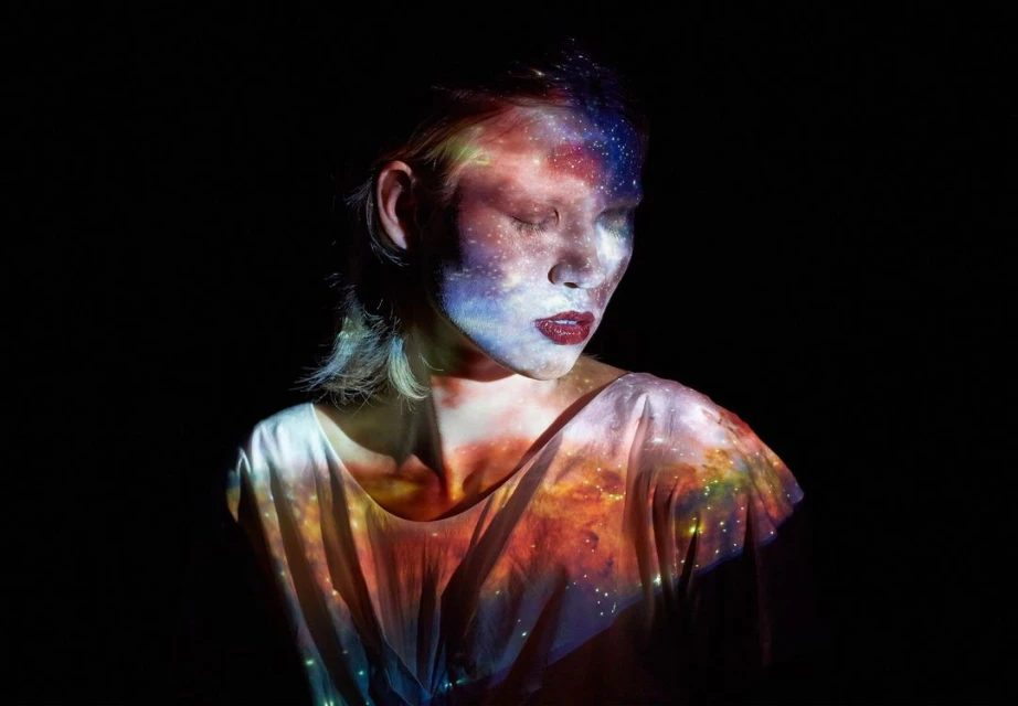 Artistic portrait of a young woman with galaxy projection on her face and body, wearing a white shirt against a dark background.