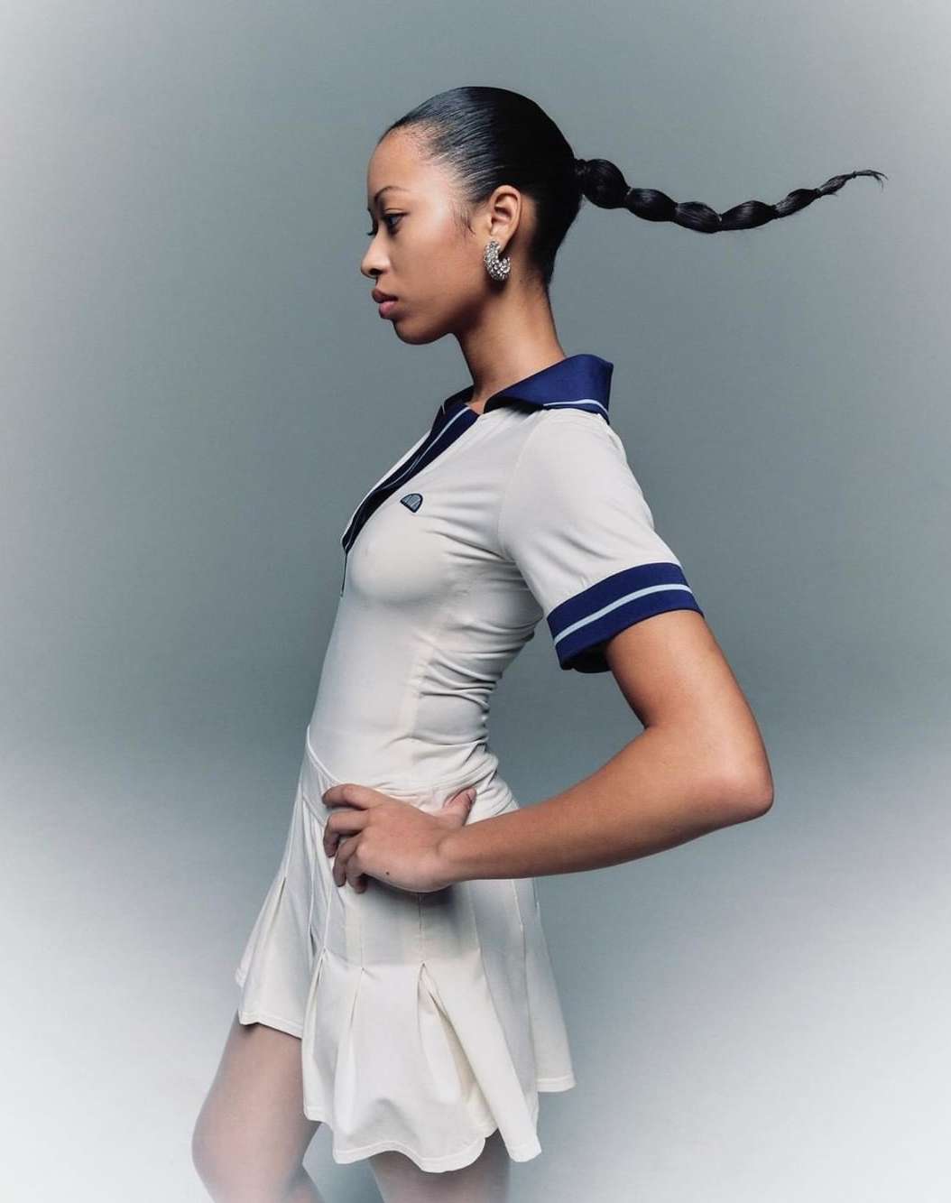 Young woman in a tennis outfit poses in a studio. She has a high ponytail with a unique twist, with her hand on her hip.
