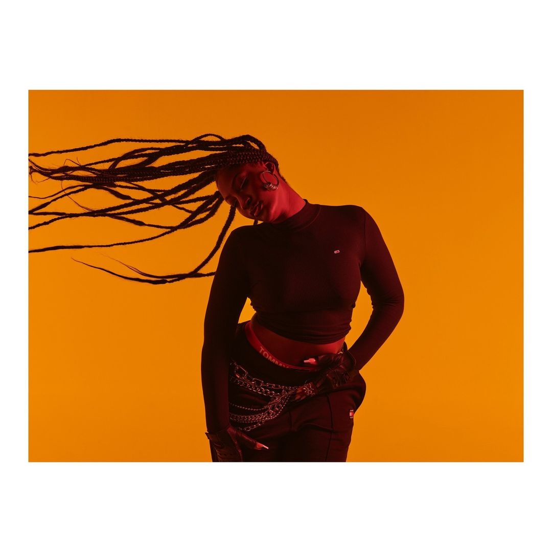 Studio shot of a woman with long braids wearing a black long-sleeved shirt and black pants against a solid orange background.